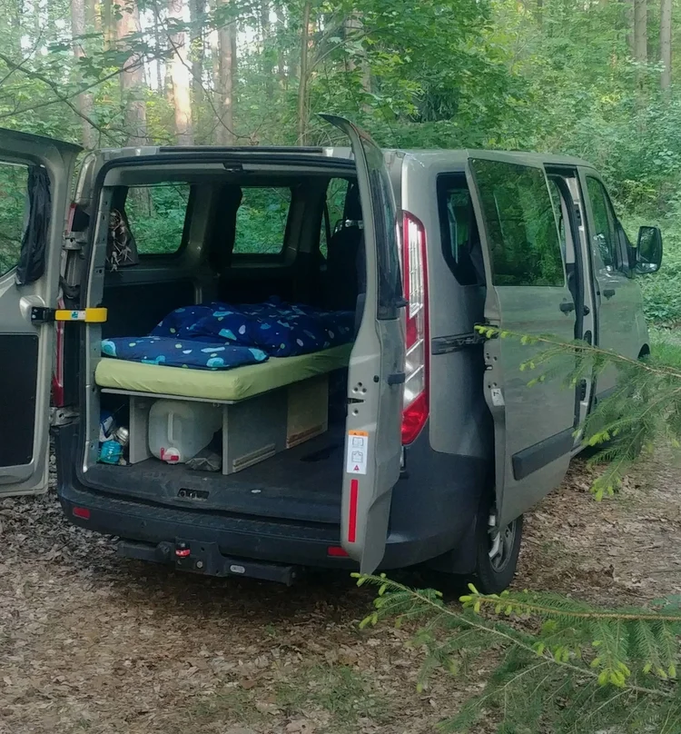 Grauer Ford Transit Custom im Wald mit ausgebautem Bett und Stauraum im Heck