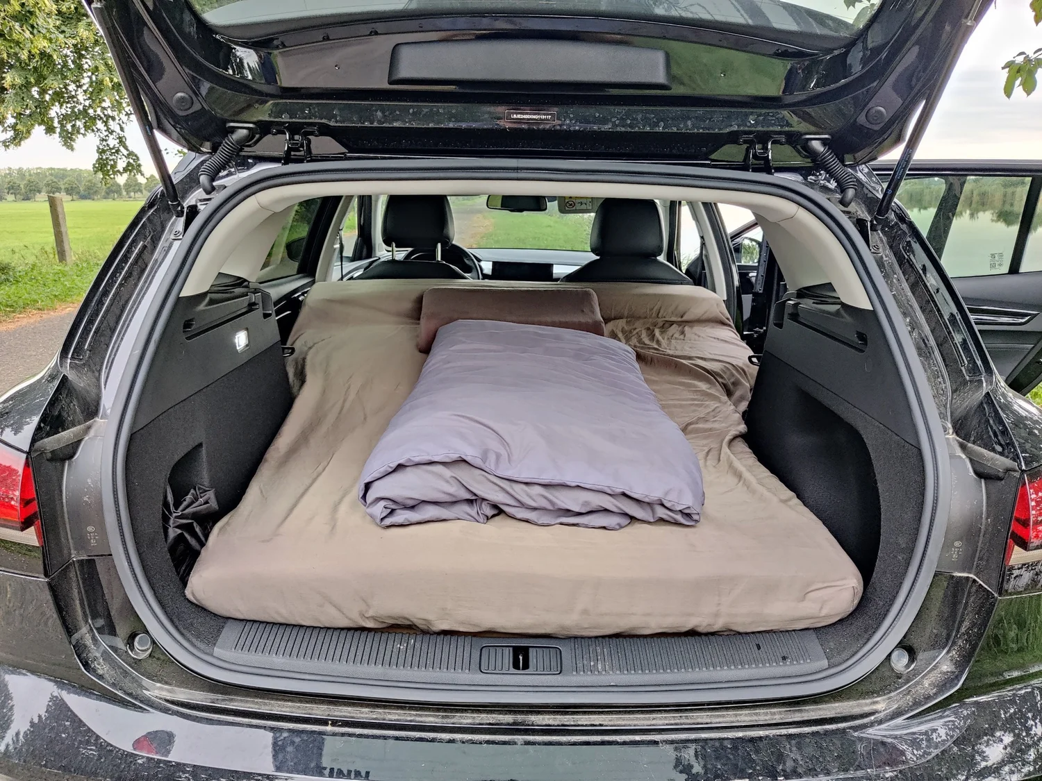 Gemachtes Bett mit Decke und Kissen im Kofferraum eines schwarzen Kombi, geparkt am Seeufer